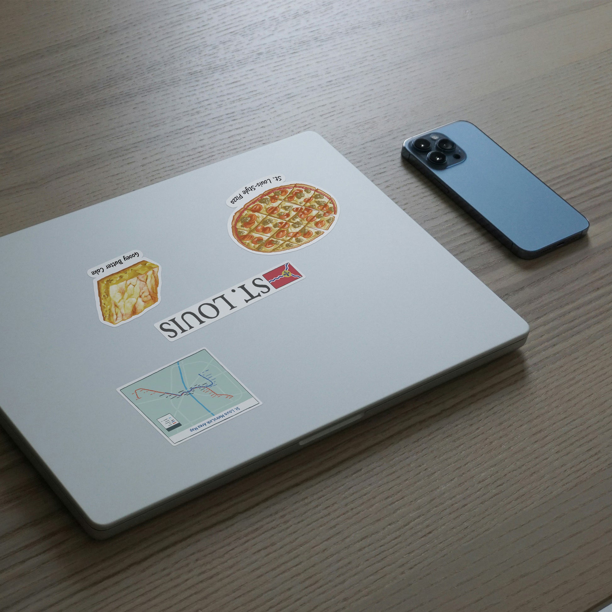 White laptop lid with St. Louis stickers: a ST. LOUIS wordmark, St. Louis-style pizza, gooey butter cake and a MetroLink map.