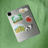 A silver tablet lying on a textured green fabric surface, decorated with four colorful illustrated Panama stickers and accompanied by white wireless earbuds.