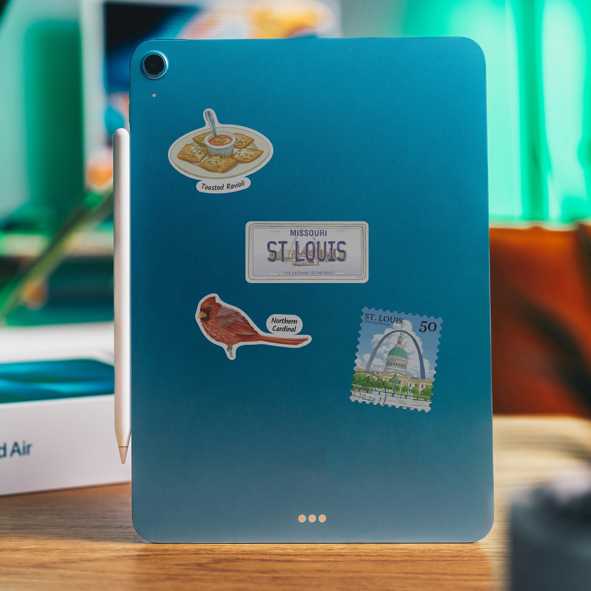 Teal tablet back with St. Louis stickers: toasted ravioli, a Missouri licence plate and a northern cardinal.
