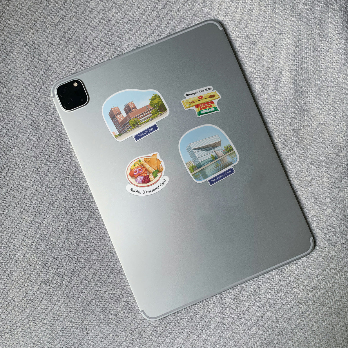 A silver tablet lying on a textured gray fabric surface, decorated with four colorful Oslo stickers featuring local landmarks and food illustrations