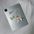 A silver tablet lying on a textured gray fabric surface, decorated with four colorful Atlanta travel-themed stickers and accompanied by white wireless earbuds.