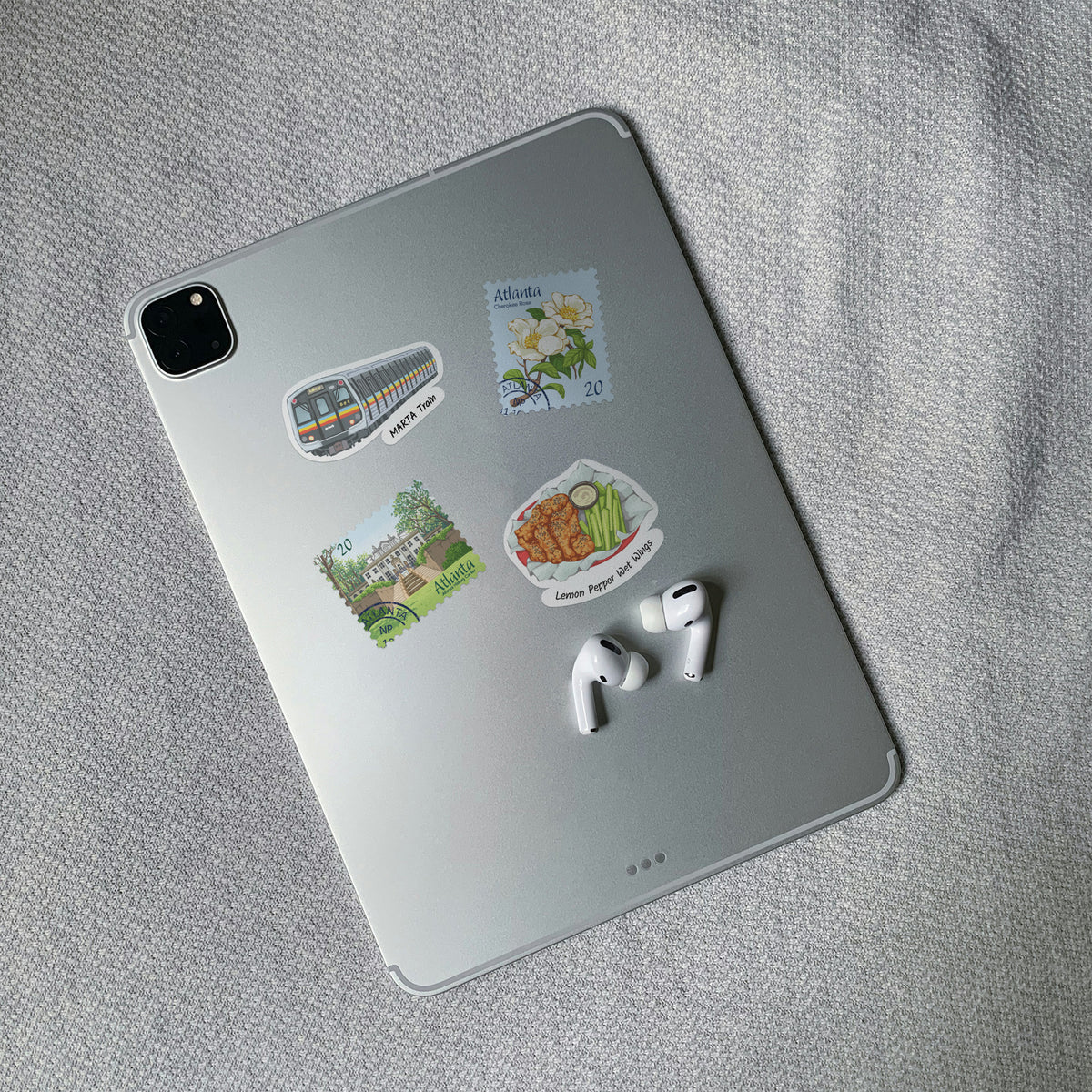 A silver tablet lying on a textured gray fabric surface, decorated with four colorful Atlanta travel-themed stickers and accompanied by white wireless earbuds.