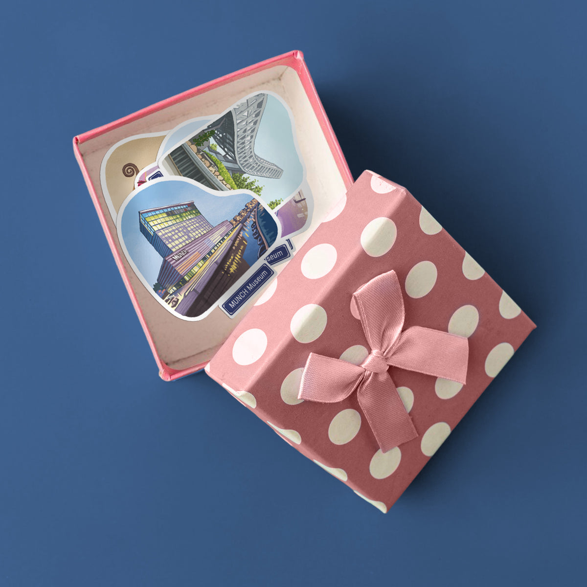 A pink gift box with white polka dots and a satin bow, shown open with two heart-shaped souvenir stickers inside. The stickers include MUNCH Museum, and the box is set against a solid blue background.