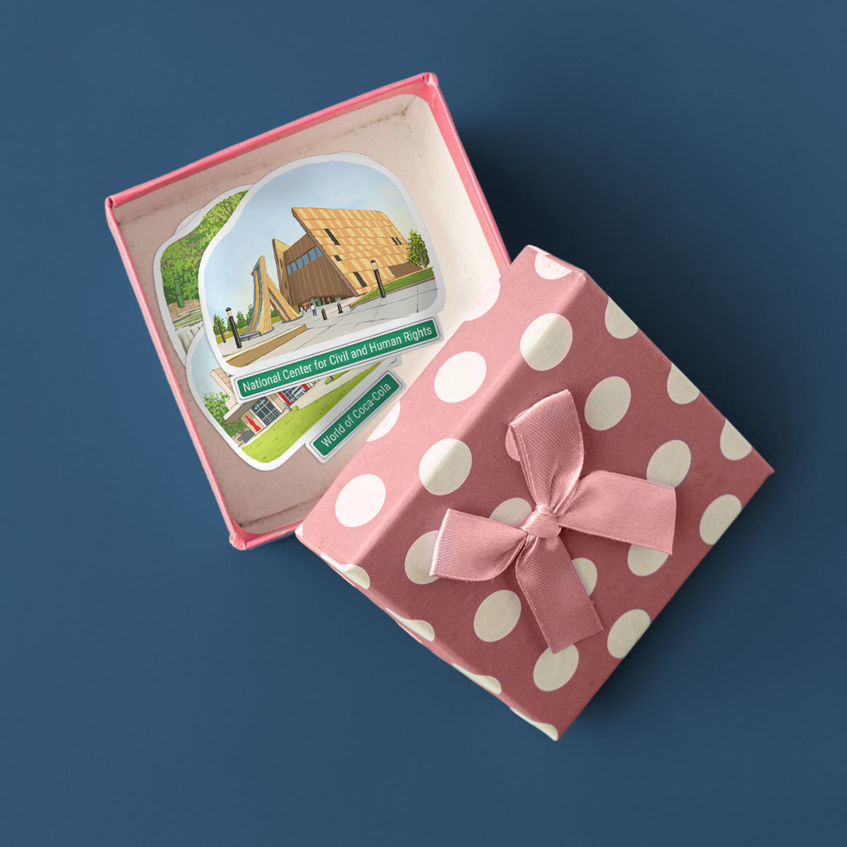 A pink gift box with white polka dots and a matching ribbon bow, shown open on a dark blue background. Inside the lid is a small illustrated card featuring a building and the text 'National Center for Civil and Human Rights' and 'World of Coca-Cola'