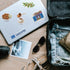 White laptop beside a packed suitcase, with 'Santorini' lettering, olive branch and Greek coffee stickers