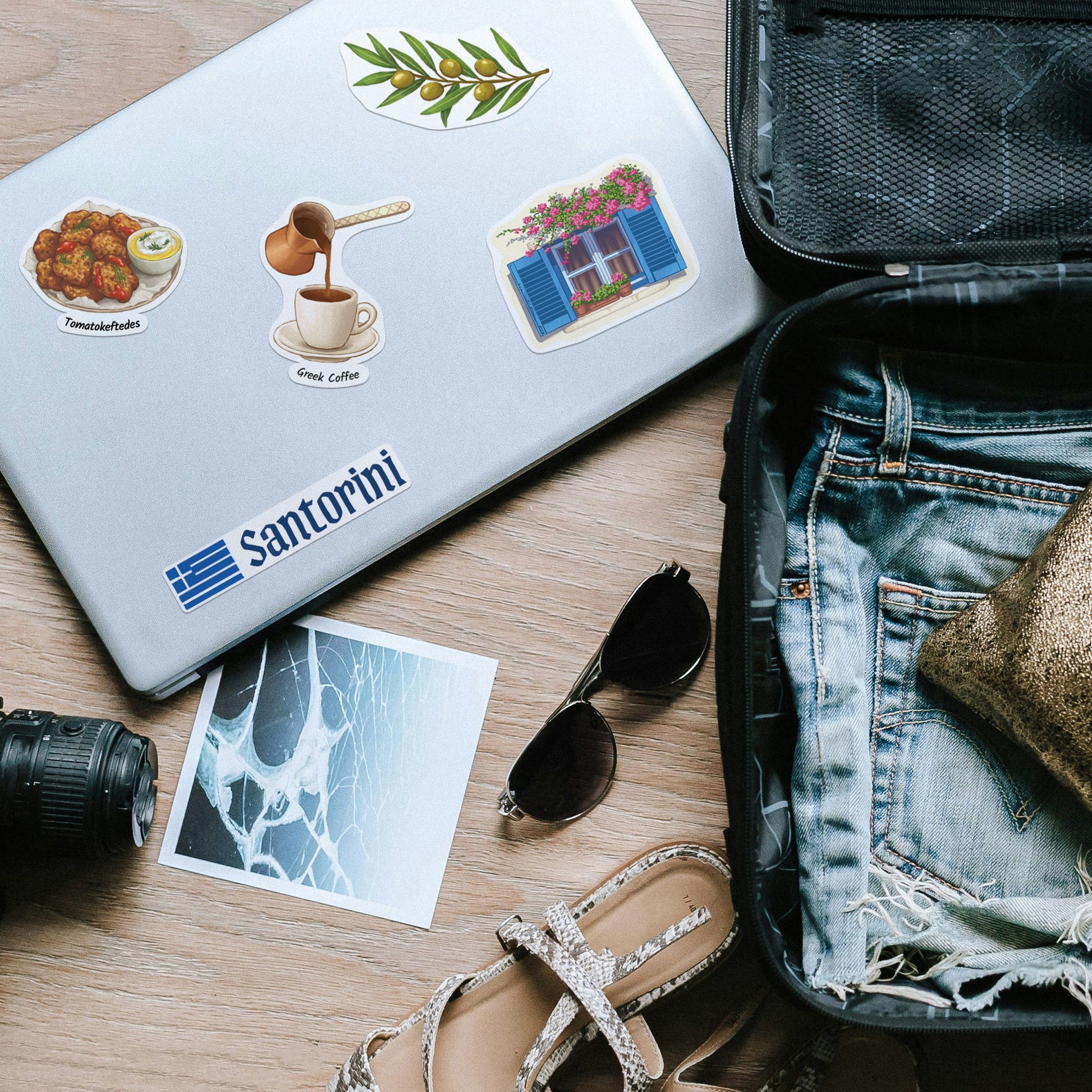 White laptop beside a packed suitcase, with 'Santorini' lettering, olive branch and Greek coffee stickers