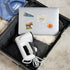 A silver laptop with colorful stickers, including a moose and an Oslo transit map, sits inside an open suitcase along with a white handheld heat gun and folded clothing on a beige textured rug.