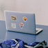 A silver laptop on a blue table with several colorful stickers on its lid, including a map, a food illustration, and a The Varsity Sign graphic. A blue mesh bag and sunglasses are partially visible in the foreground.