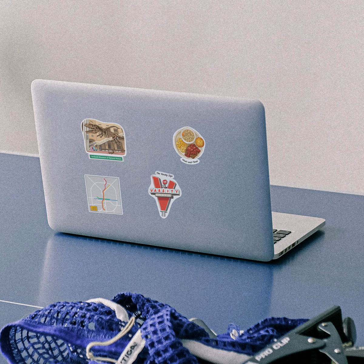 A silver laptop on a blue table with several colorful stickers on its lid, including a map, a food illustration, and a The Varsity Sign graphic. A blue mesh bag and sunglasses are partially visible in the foreground.