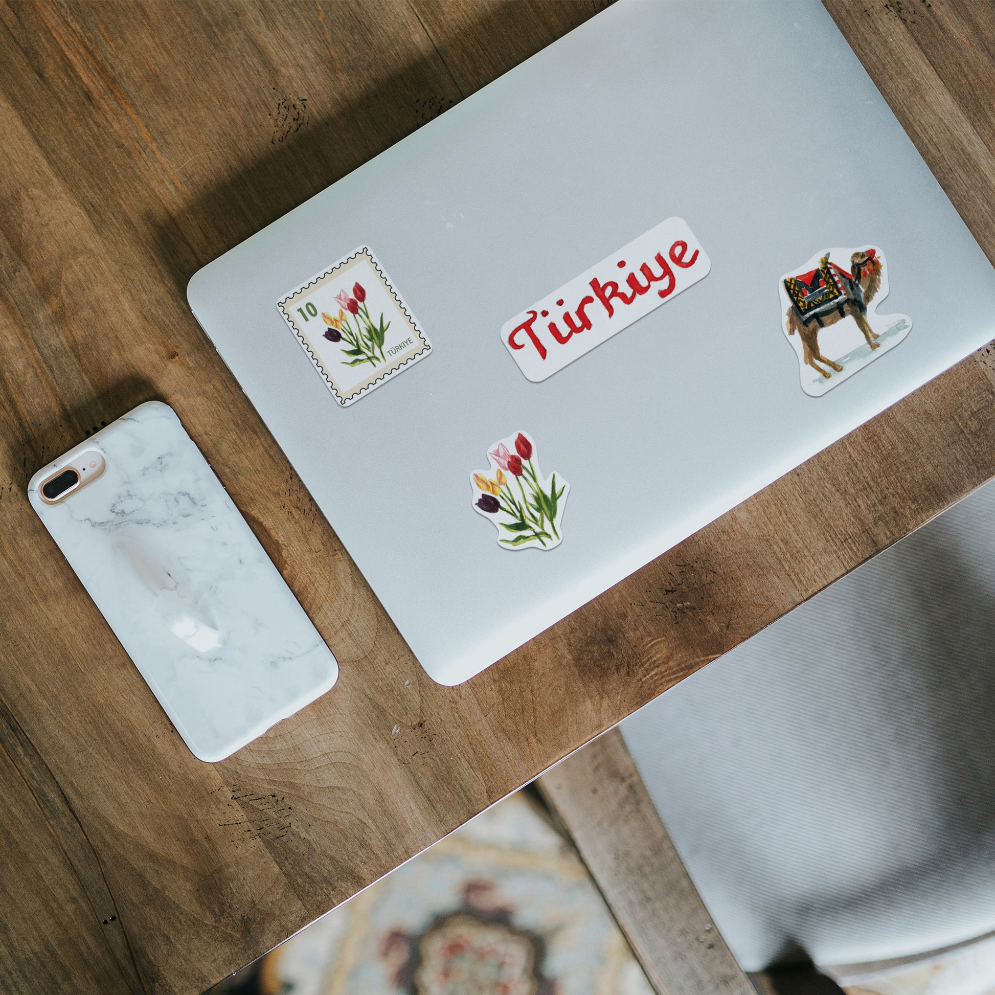 "Turkiye", tulip stamp and camel stickers on a silver laptop lid beside a marble phone case