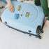 A light blue hard-shell suitcase with four wheels and a telescoping handle, being held by two people on a white floor. The suitcase has three decorative Atlanta travel stickers on the lid, including one that says 'ATLANTA'.