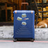 A blue hard-shell suitcase with a telescoping handle and four wheels, decorated with three Oslo travel stickers, standing on a street curb with a dog lying in the background.