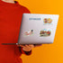 A person in a red ribbed sweater holds a silver laptop against a bright orange background. The laptop lid has several stickers, including a blue 'COPENHAGEN' text sticker and illustrated travel-themed stickers.