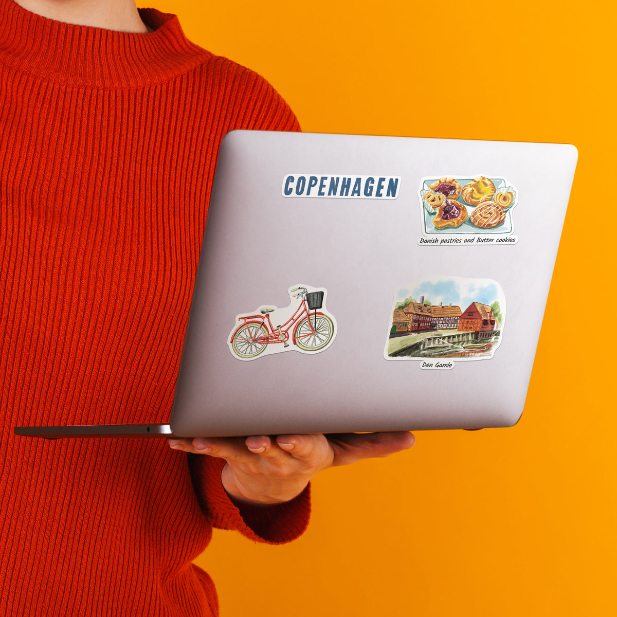 A person in a red ribbed sweater holds a silver laptop against a bright orange background. The laptop lid has several stickers, including a blue 'COPENHAGEN' text sticker and illustrated travel-themed stickers.