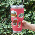 A hand holds a pink insulated travel tumbler with a clear lid and metal straw, decorated with four colorful architectural and cultural stickers, against a blurred green hedge background.