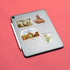 A silver tablet lying on a coral-pink background, decorated with four travel-themed stickers including 'The Blind Tower,' 'The Little Mermaid,' 'Yacht Clubbers,' and a 'Danmark 20' flower stamp. A white stylus rests beside the tablet.