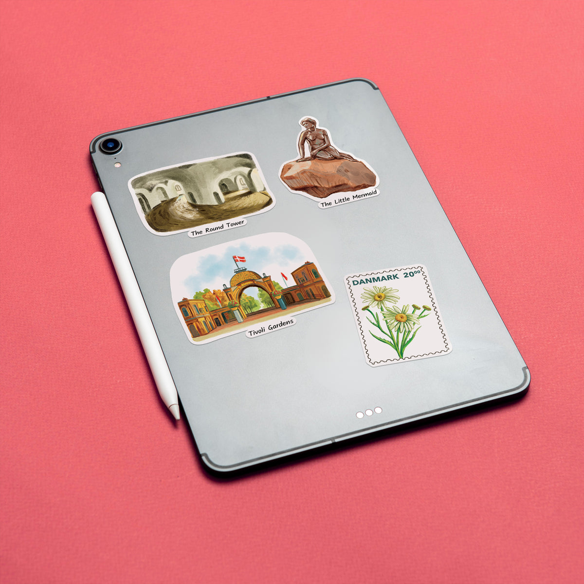 A silver tablet lying on a coral-pink background, decorated with four travel-themed stickers including 'The Blind Tower,' 'The Little Mermaid,' 'Yacht Clubbers,' and a 'Danmark 20' flower stamp. A white stylus rests beside the tablet.