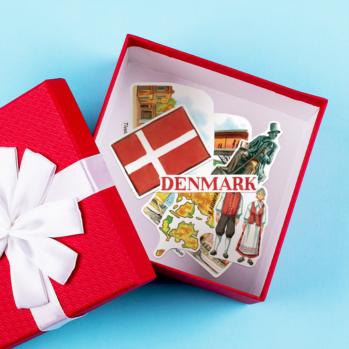 A red gift box with a white ribbon is open to reveal a set of Denmark-themed stickers, including the Danish flag, a map of Denmark, traditional figures, and landmarks, on a light blue background.