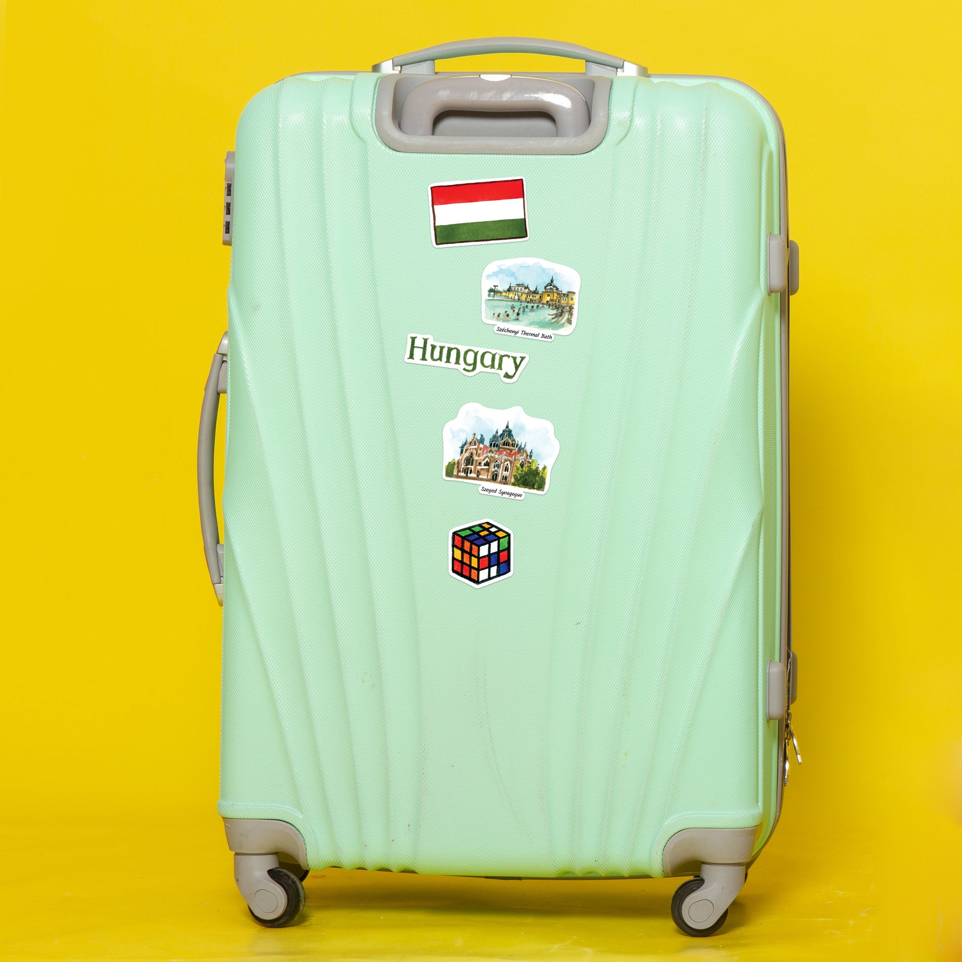 Hungarian flag, 'Hungary' label, Rubik's cube and landmark stickers on a mint suitcase against yellow