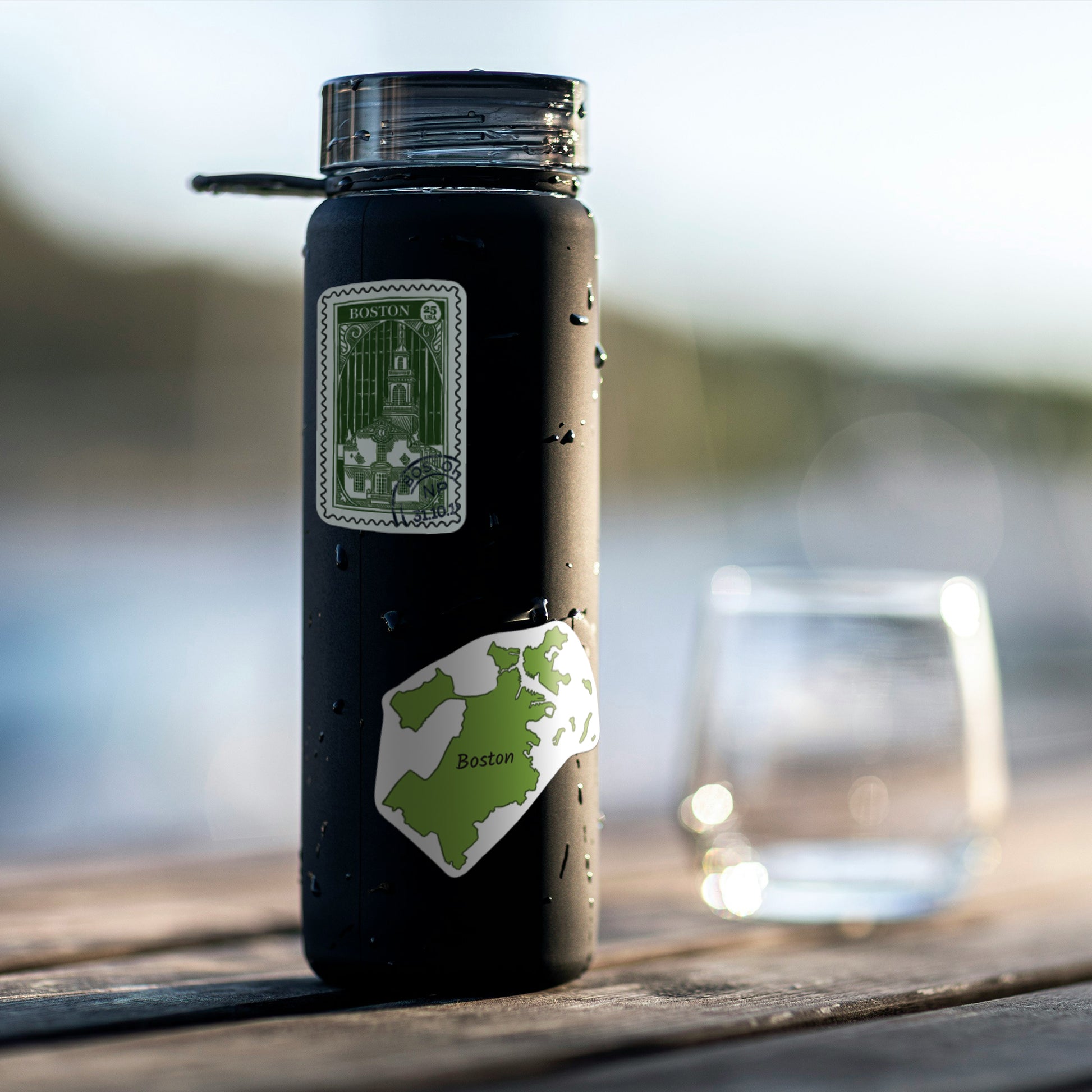 A Boston stamp sticker and a green state map sticker on a black water bottle beaded with water