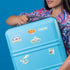 A person holding a light blue hard-shell suitcase decorated with various Detroit-themed stickers, set against a solid blue background.