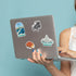 A person holds a silver laptop decorated with several die-cut travel stickers, including Ambassador Bridge, a vintage car, a Detroit-style pizza slice, and Belle Isle Fountain, against a solid light blue background.