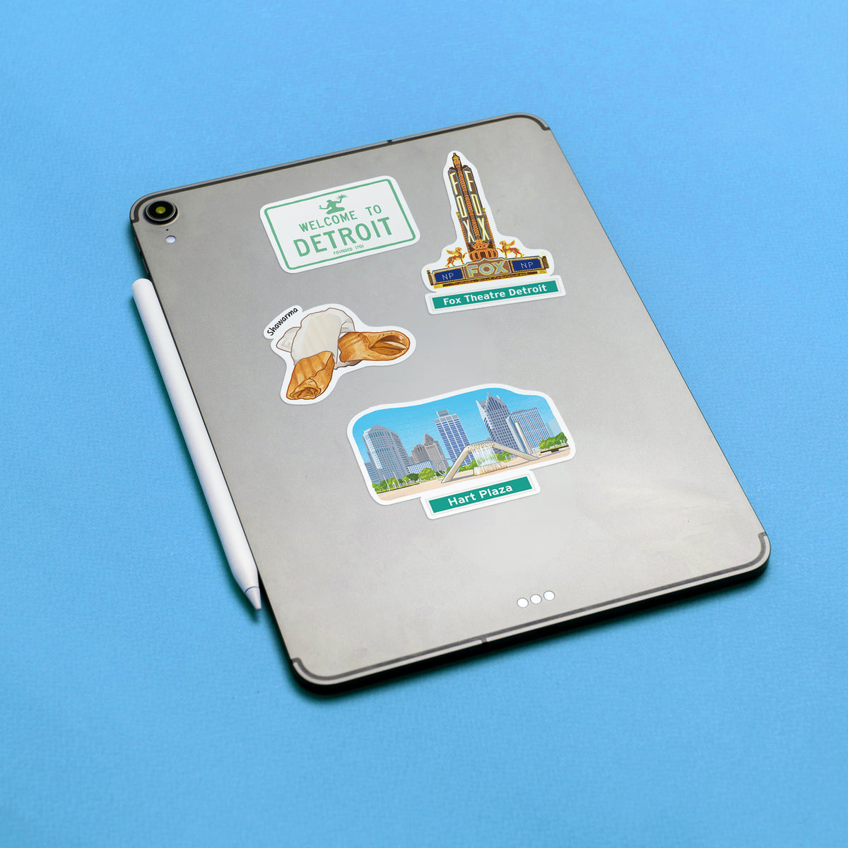 A silver tablet on a bright blue background, decorated with four Detroit-themed stickers including a 'Welcome to Detroit' sign, a Fox Theatre Detroit sticker, a Shawarma sticker, and a Hart Plaza sticker. A white stylus lies beside the tablet.