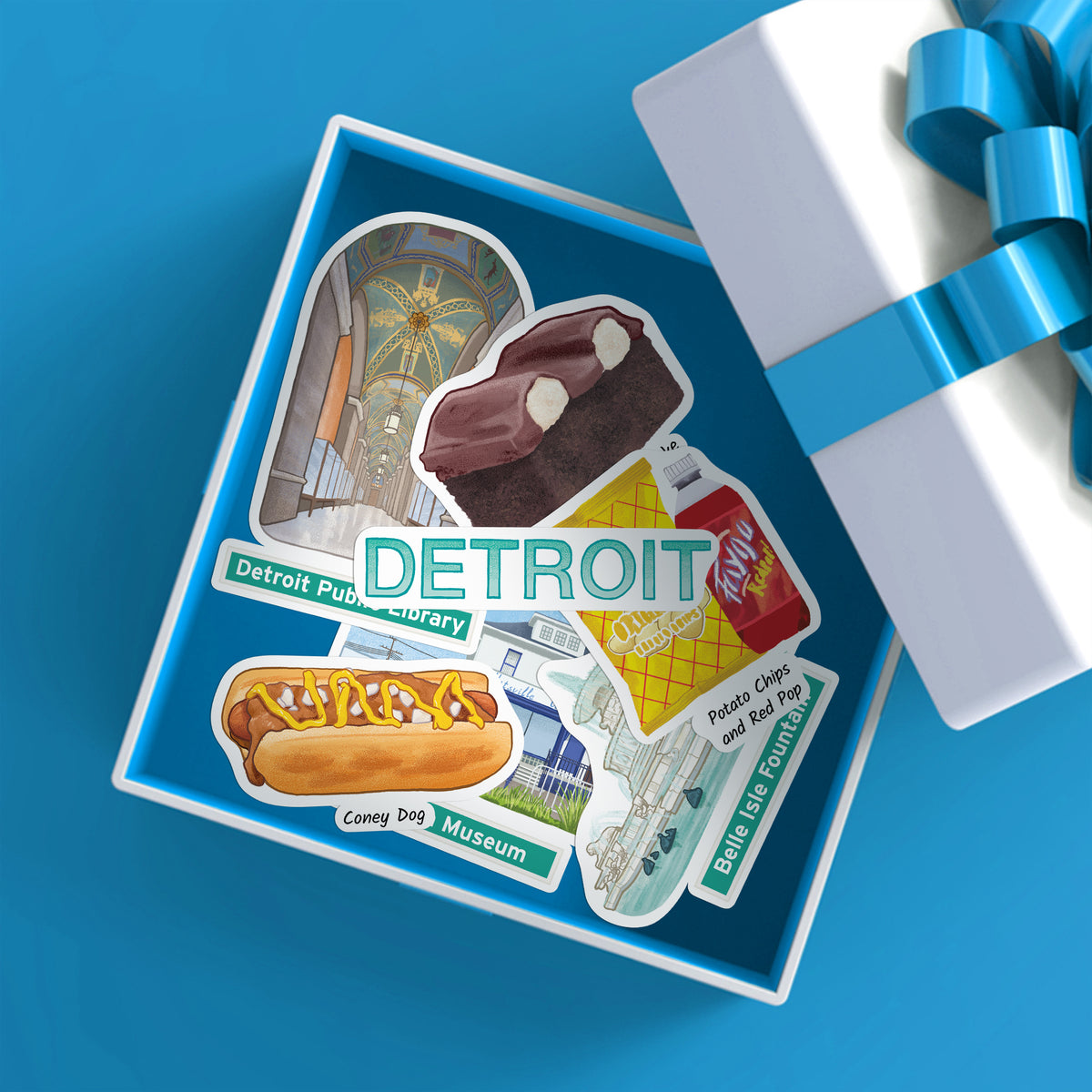 A square gift box with a blue interior displays a Detroit-themed sticker collage featuring the Detroit Public Library, Coney Dog Museum, a Red Pop soda bottle, and other local illustrations. A white gift box with a blue ribbon sits beside it on a bright blue background.