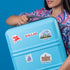 A person holding a light blue hard-shell suitcase decorated with colorful travel stickers, including a red Dallas logo and a purple flower stamp, against a solid blue background.