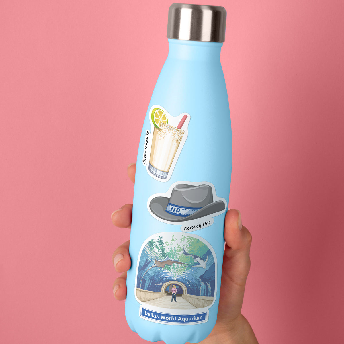 A light blue insulated water bottle with a stainless steel cap, held in a hand against a solid pink background. The bottle features three style stickers: a Frozen Margarita, a gray cowboy hat with 'Cowboy hat' text, and an illustration of the Dallas World Aquarium.