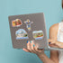 A person holds a silver laptop decorated with four colorful die-cut stickers, including a Texas BBQ sticker, a Big Tex sticker, and two landmark stickers, against a solid light blue background.