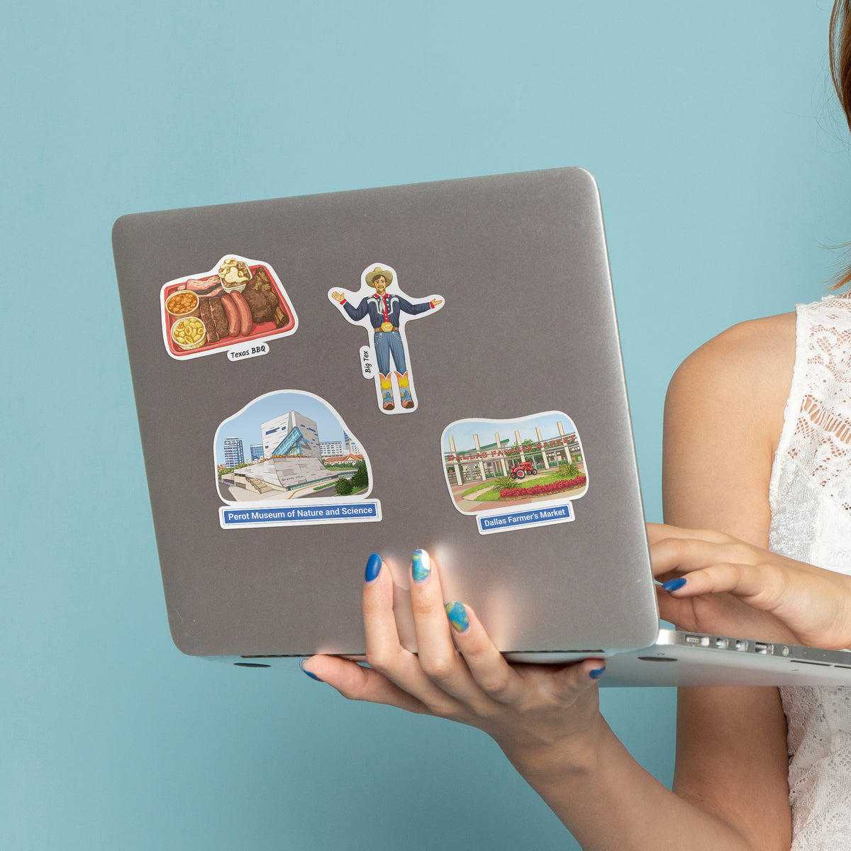 A person holds a silver laptop decorated with four colorful die-cut stickers, including a Texas BBQ sticker, a Big Tex sticker, and two landmark stickers, against a solid light blue background.
