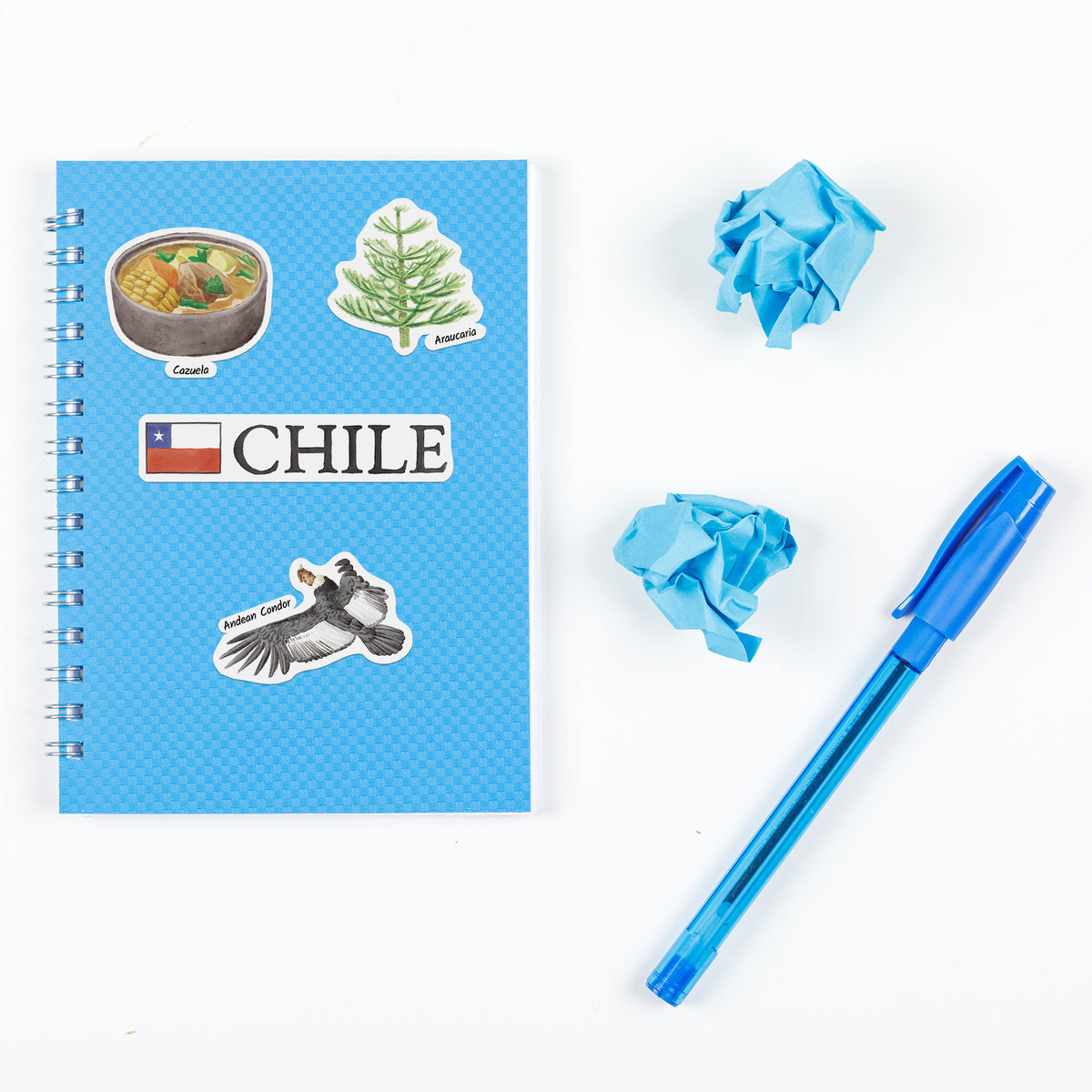 A spiral-bound notebook with a blue cover decorated with Chile-themed stickers, shown on a white background alongside two crumpled blue paper balls and a blue pen.