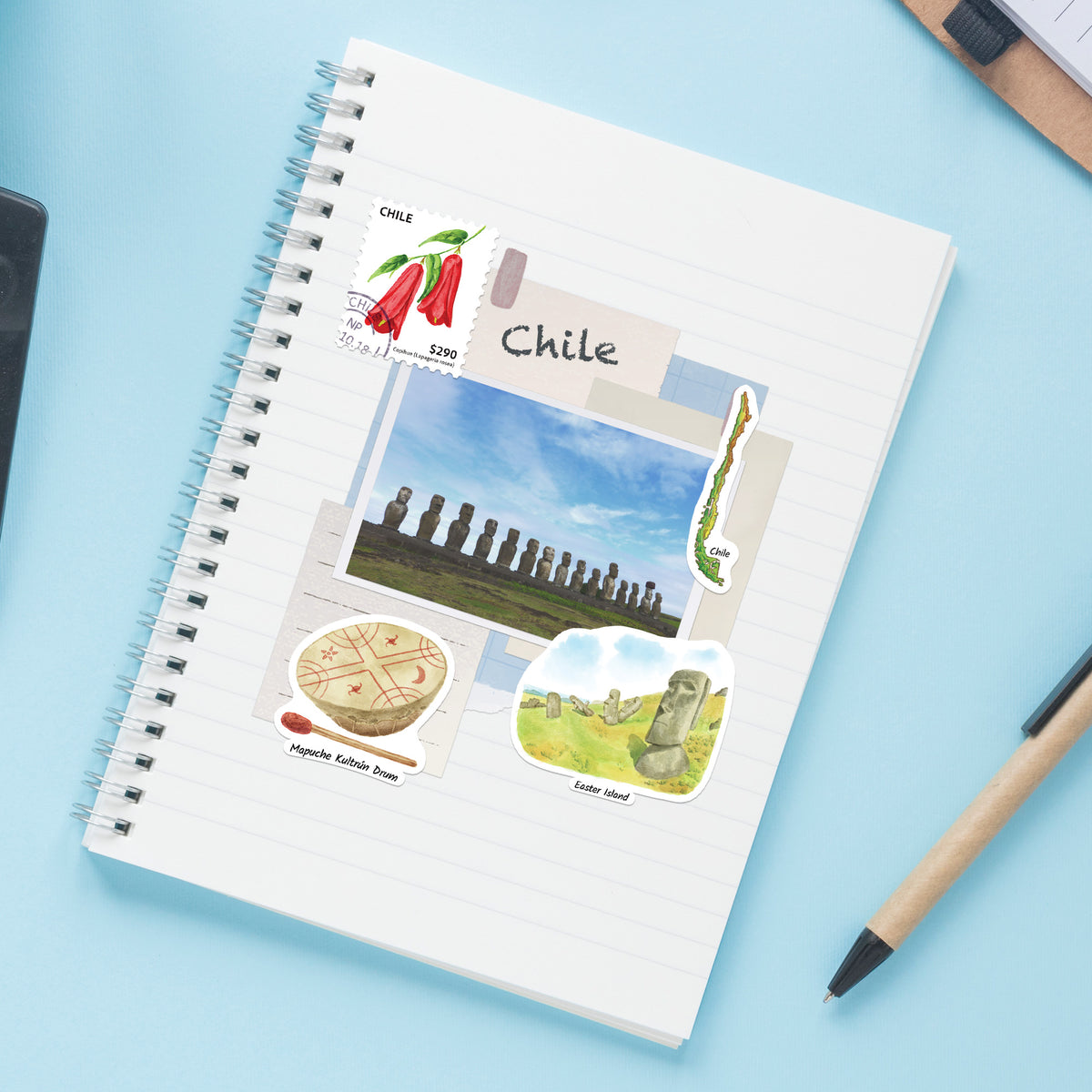 A spiral-bound notebook with Chile-themed stickers and illustrations, photographed from above on a light blue surface with a pen nearby.