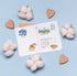 A white postcard with colorful illustrated stamps and handwritten address details, arranged on a light blue background with cotton bolls and wooden heart cutouts around it.