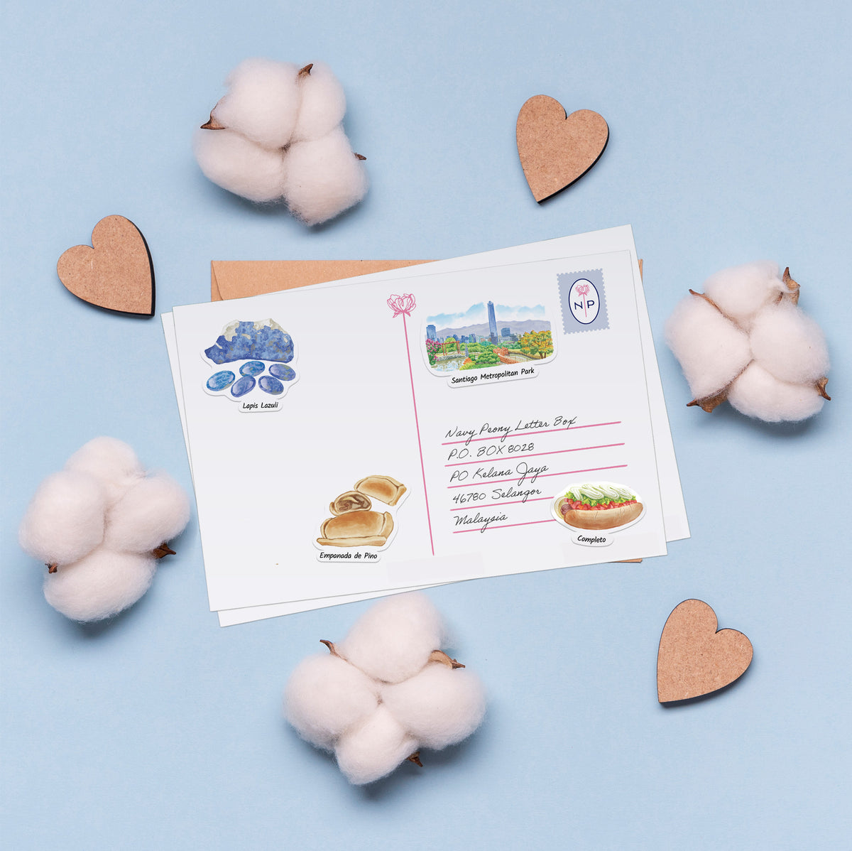 A white postcard with colorful illustrated stamps and handwritten address details, arranged on a light blue background with cotton bolls and wooden heart cutouts around it.