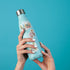 A light blue insulated water bottle with a stainless steel cap, held by two hands against a solid turquoise background. The bottle is decorated with three colorful travel-themed stickers.