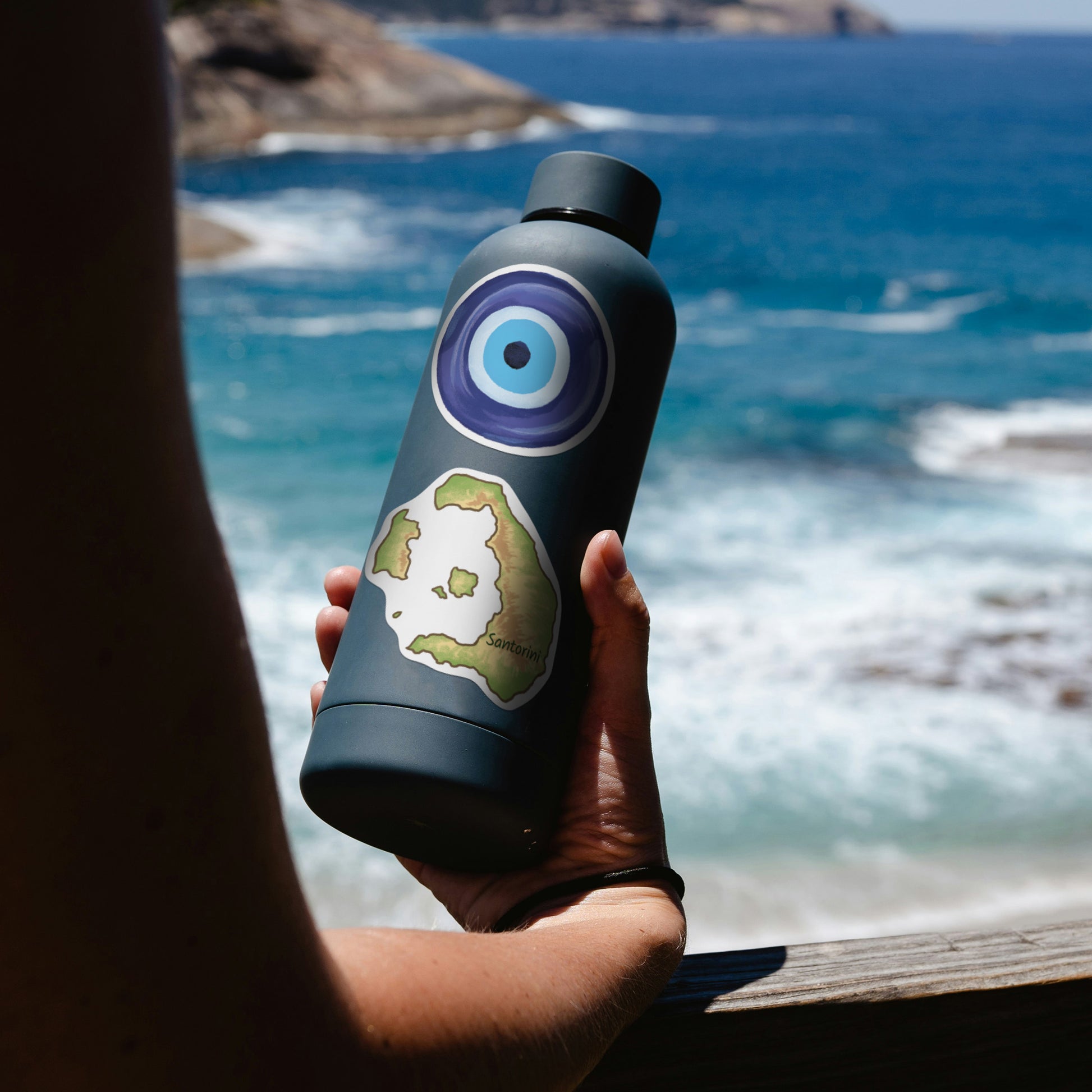 Hand holding a navy water bottle by the sea, with evil eye charm and Santorini island map stickers