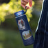 A navy blue insulated water bottle with custom illustrations of a couple and Oslo Cathedral, held by a hand against a blurred outdoor background.