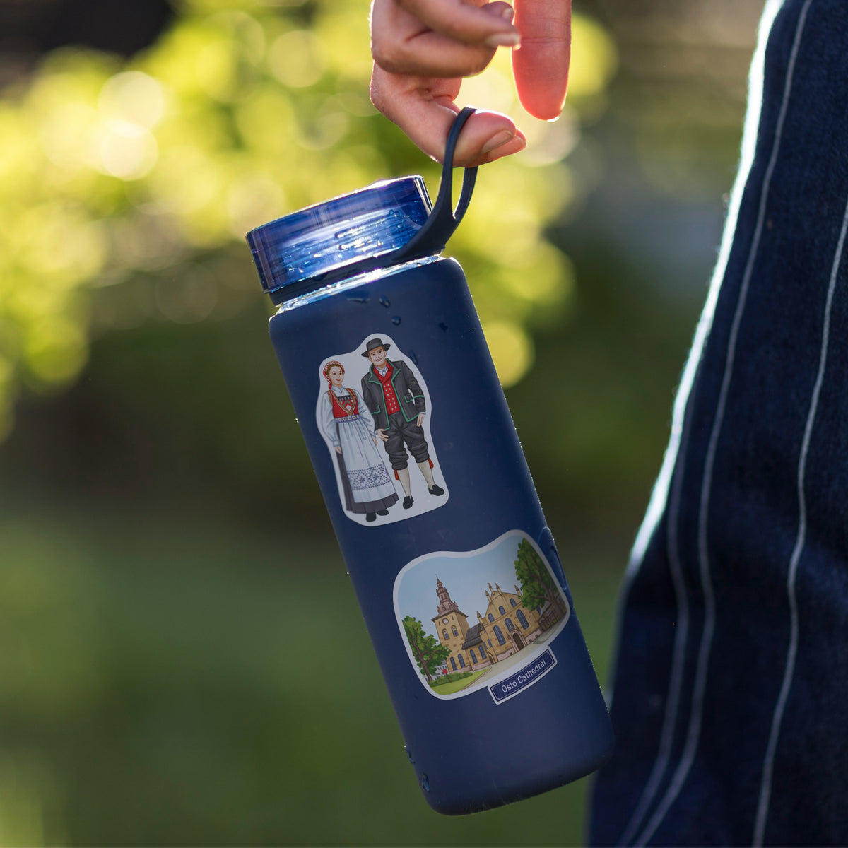 A navy blue insulated water bottle with custom illustrations of a couple and Oslo Cathedral, held by a hand against a blurred outdoor background.