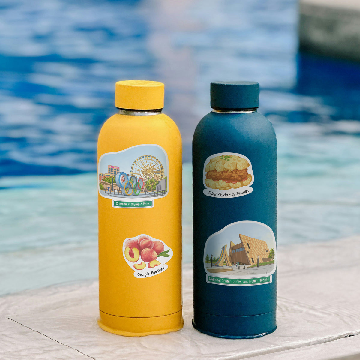 Two insulated stainless steel water bottles standing side by side on a stone ledge with a blurred swimming pool in the background. The bottle on the left is mustard yellow with a yellow cap and a colorful Georgia Peaches sticker, and the bottle on the right is dark teal-blue with a dark cap and a Fried Chicken & Biscuits sticker.